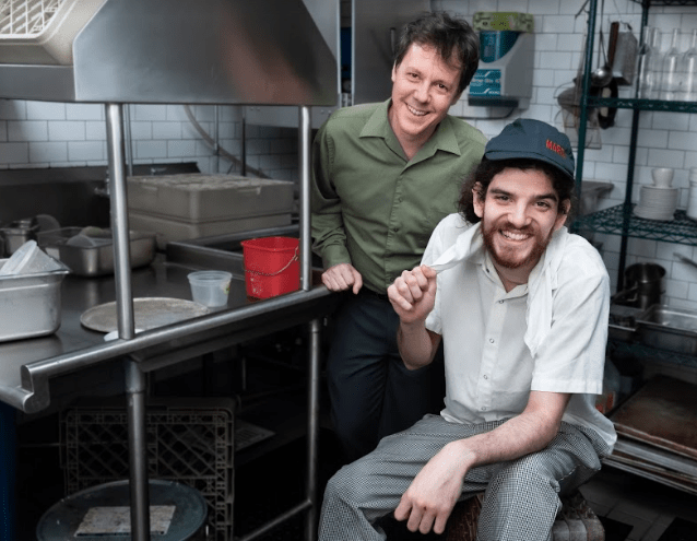 jake-and-tim A young man with a goatee is smiling next to his employment specialist, who is also smiling, they are in a restaurant kitchen.