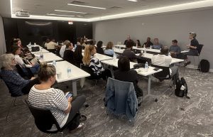 A conference room with tables and about 20 attendees listening to 5 speakers, including Wendy Banner on matters related to customized employment.